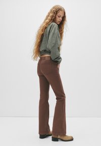 Jeans maro evazați cu talie înaltă și buzunare pe spate, asortați cu o cămașă lungă cu mâneci lungi în carouri verzi. Modelul poartă cizme scurte de culoare bej.