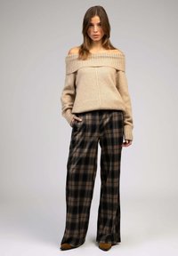 Pull en tricot beige avec design dénudé sur les épaules et accents en côtes, associé à un pantalon à carreaux large noir et beige. Bottines en cuir suédé.