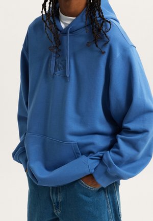 Personne portant un sweat à capuche bleu Vans avec une poche avant et un jean bleu, mains dans les poches, sur un fond clair uni.