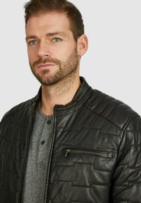 Svart läderbombarjacka med ett quiltat mönster, zip-ficka på bröstet och ståkrage. Buren över en grå henley-tröja.