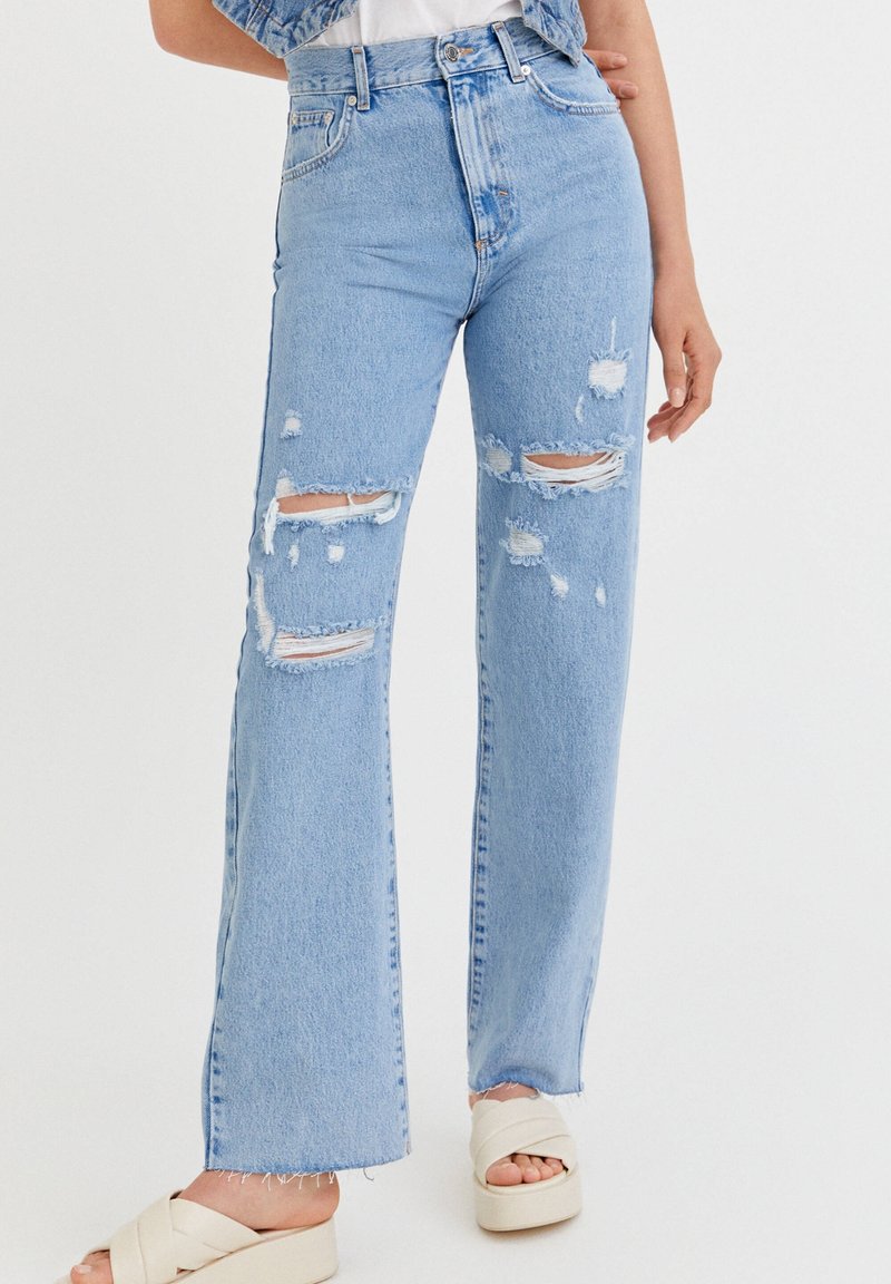 Femme portant un jean déchiré taille haute bleu clair et des sandales à plateforme blanches, debout, une main détendue le long du corps.