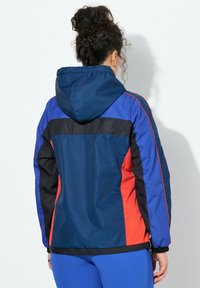 Barevná bunda kombinující tmavě modré, červené a černé panely, s kapucí a elastickými manžetami. Texturovaná látka přidává designu hloubku.