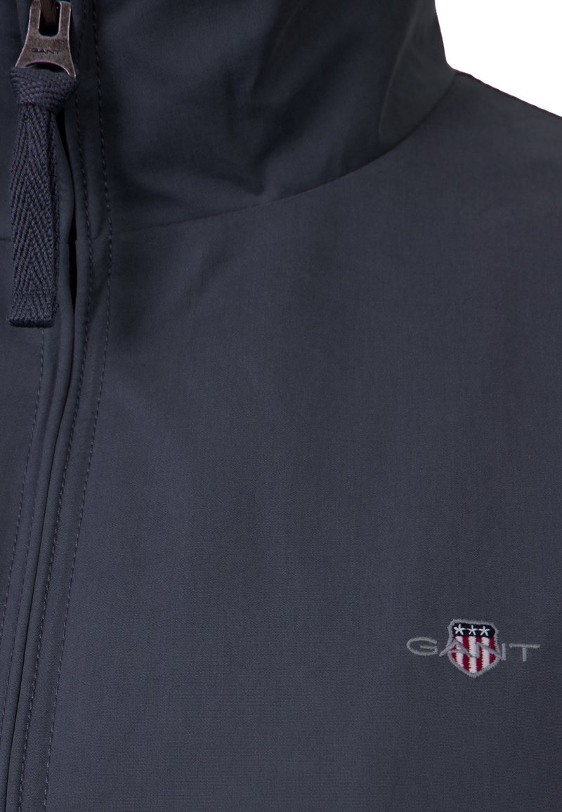 Primo piano della cerniera di una giacca blu scuro con tiretto intrecciato e logo GANT ricamato che presenta uno scudo con stelle e strisce.