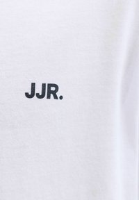 Λευκό ύφασμα με μαύρο κείμενο "JJR." τυπωμένο, που δείχνει την υφή του υφάσματος και καθαρά, έντονα γράμματα.