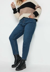 Pletený svetr s černými, béžovými a světle hnědými pruhy; v kombinaci s tmavě modrými džínami a černými šněrovacími botami s kovovými detaily.