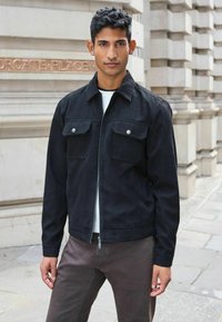 Schwarze Wildlederjacke mit Reißverschluss, zwei Brusttaschen und spitzem Kragen. Über einem hellen, strukturierten Pullover getragen, kombiniert mit dunklen Hosen.