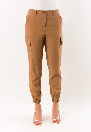 Pantaloni cargo marroni con polsini elastici indossati con sandali beige con tacco alto su sfondo chiaro.