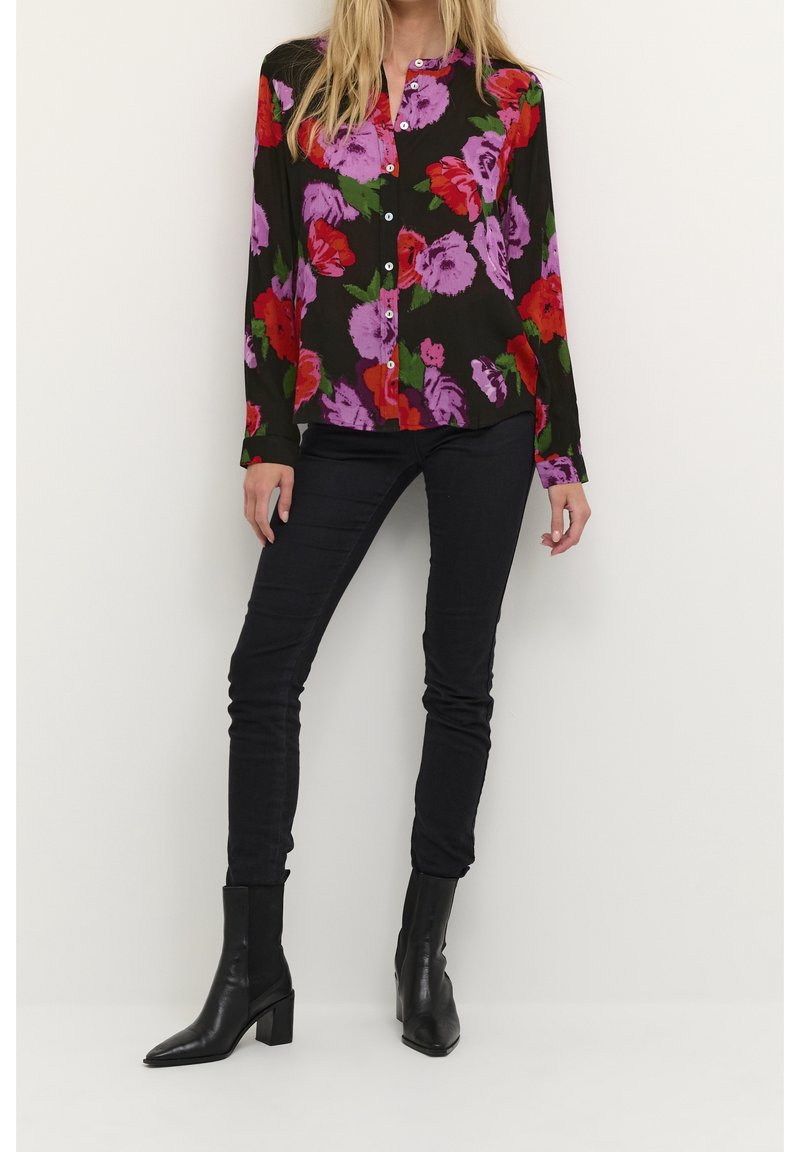 Femme portant un chemisier noir à fleurs avec des boutons, un pantalon noir ajusté et des bottines à talons noires, debout contre un mur blanc uni.