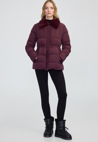 Giovane donna che indossa un giubbino imbottito bordeaux con colletto in pelliccia sintetica, leggins neri e stivali invernali foderati in pelliccia nera, in piedi contro uno sfondo neutro.