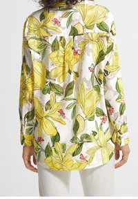 Blouse à manches longues en tissu léger, ornée d'un motif floral avec des fleurs jaunes et des feuilles vertes. Vue de dos montrant un détail de pli.