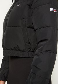 Sort cropped pufferjakke med en shiny overflade, elastisk bund og lynlås foran. Har logodetalje på skulderen og en lappet lomme.