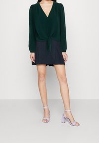 Blusa verde scuro con scollatura a V e chiusura a bottoni, legata in vita. Gonna mini nera con pieghe. Sandali con tacco a blocco color lavanda.