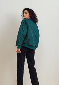 Teal bomberjakke med puffet tekstur, ribbet halsudskæring og manchetter, parret med sorte bukser, vist fra ryggen mod en ensfarvet baggrund.