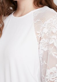 Vit topp med en slät tygkropp och genomskinliga spetsärmar med blommönster, som skapar en texturerad kontrast i designen.