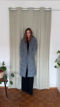 Długi szary puchowy płaszcz z kapturem, wyposażony w boczne kieszenie i luźny krój. Płaszcz ma gładką fakturę i jest noszony na czarnych spodniach.