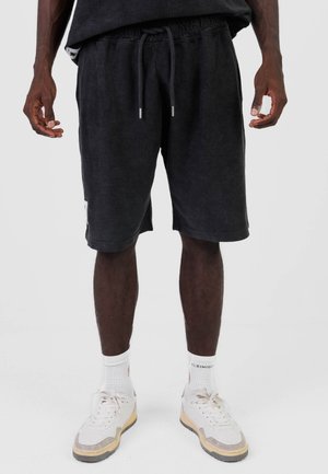 Mężczyzna w czarnych prążkowanych szortach ze sznurkiem, białych skarpetkach i białych trampkach, stojący na jednolitym tle.