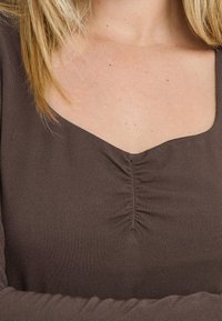 Top marrone a coste con scollatura a V, caratterizzato da un dettaglio arricciato al centro. Realizzato in un materiale morbido, con un design aderente.