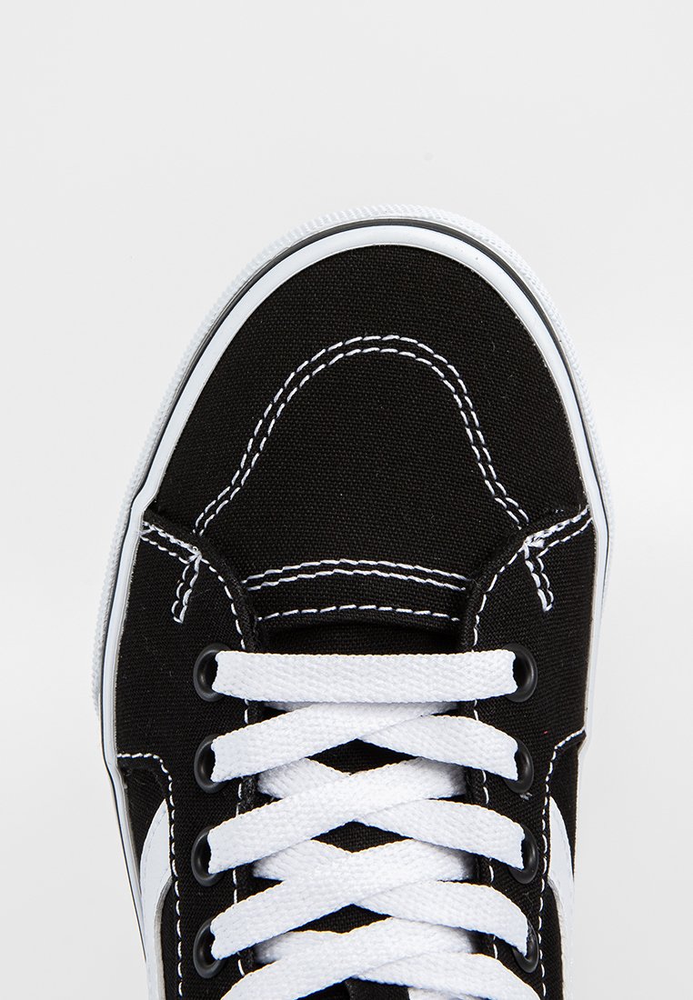 Zapatilla de lona negra con cordones blancos. Presenta costuras blancas en contraste y suela de goma. Diseñada con una punta redondeada y acentos laterales.