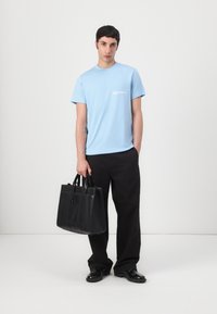 T-shirt en coton bleu clair avec des manches courtes et un logo blanc, associé à un pantalon noir ample et un sac fourre-tout en cuir noir.