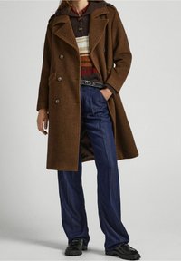 Cappotto marrone a spina di pesce con design doppiopetto, grandi bottoni e due tasche frontali, indossato sopra un maglione a righe multicolori e jeans a vita alta.