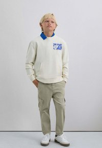 Sudadera blanca con cuello azul y logo, combinada con pantalones cargo verde claro. Zapatillas blancas con detalles grises completan el look.