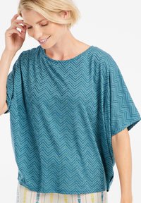 Turkio spalvos palaidinė su ševrono raštu, trumpomis rankovėmis ir laisvu siluetu. Pagaminta iš minkštos medžiagos su glotnia tekstūra ir plačiu iškirpimu.