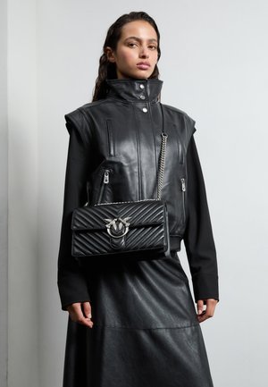 Gilet in pelle nera con collo alto, tasche con zip e una borsa a tracolla nera trapuntata con un accento in hardware argentato, indossato con una lunga gonna nera.