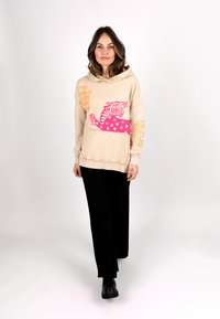 Beige hoodie with a large pink tiger graphic and stars; features a kangaroo pocket and drawstring hood. Paired with black wide-leg pants.