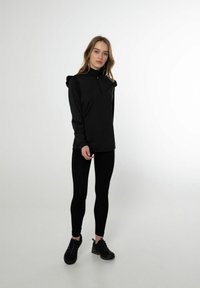Zwarte langexemplaar met ritssluiting en geraffineerde schouderaccenten, gecombineerd met zwarte leggings en zwarte sportschoenen. Glad, rekbaar materiaal.