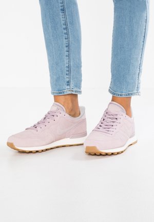 Stopy w jasno różowych butach Nike z gumowymi podeszwami i jasnoniebieskich skróconych dżinsach na białym tle.