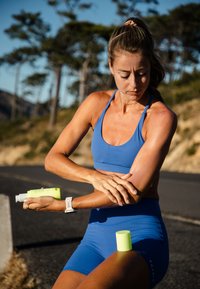 Femme en tenue de sport bleue appliquant de la crème solaire sur son bras en plein air par une journée ensoleillée, avec des arbres et une route en arrière-plan.
