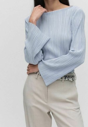 Mujer con blusa azul claro plisada de manga larga con puños acampanados y pantalones beige con cinturón de estampado de piel de serpiente.