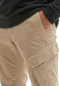 Beige Cargo-Hosen mit Seitentaschen, festem Stoff und sichtbaren Nähten, kombiniert mit einem dunklen, strukturierten Pullover.