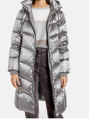 Femme portant un long manteau matelassé argenté brillant par-dessus un pull à rayures grises et blanches et un pantalon foncé, debout avec les mains détendues le long du corps.