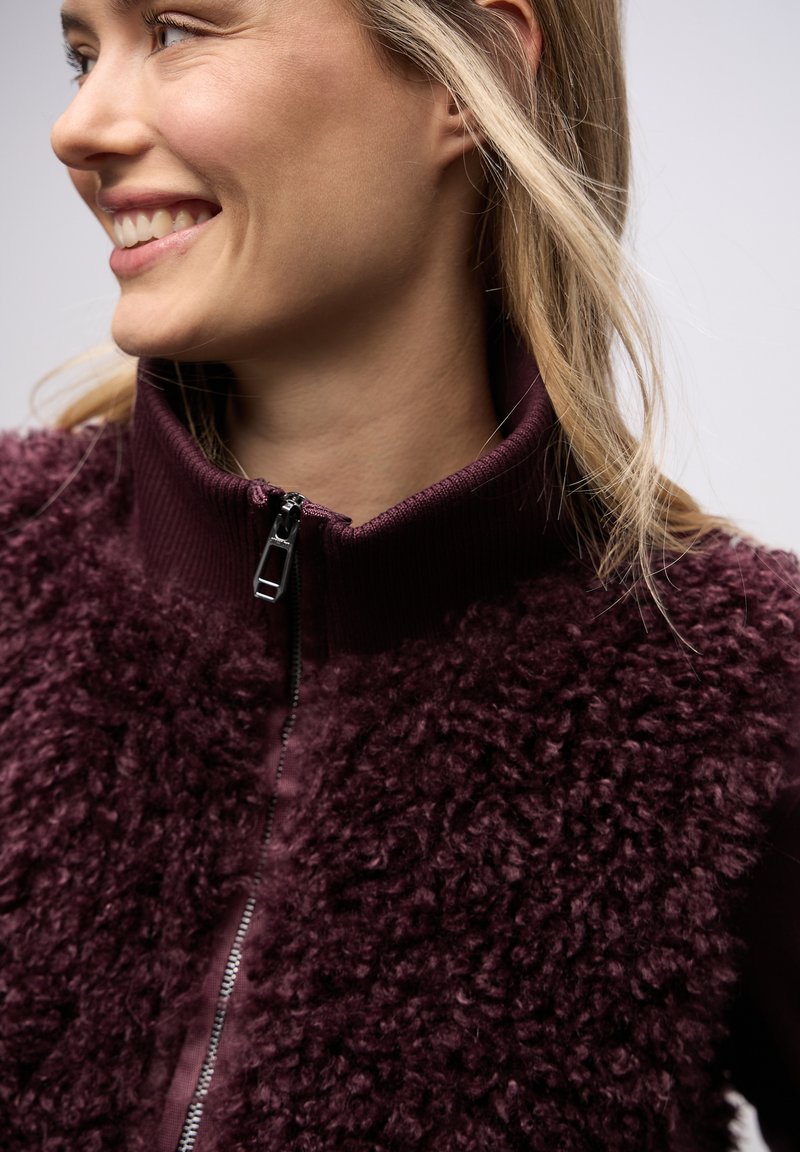 Weinrotes, zum Zuziehen geeignetes Weste mit flauschiger Textur am Körper und glattem, geripptem Kragen. Schwarzer Reißverschluss am Halsausschnitt.