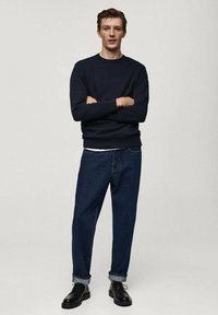 Felpa navy con colletto rotondo, abbinata a jeans in denim scuri a vestibilità rilassata e scarpe stringate nere. Design semplice, con orli arrotolati sui jeans.