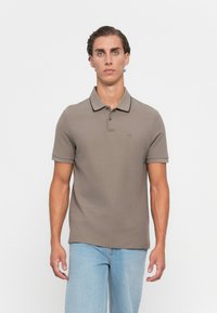 Ung mann iført taupe kortermet polo-skjorte med svart kant og lyseblå jeans, stående mot en enkel hvit bakgrunn.