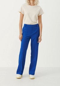 Pantaloni su misura blu con un design a gamba dritta, abbinati a una t-shirt beige chiaro. Modello che indossa sneaker bianche, in piedi davanti a uno sfondo chiaro.