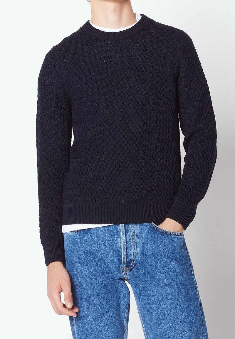 Pull en maille bleu marine avec un motif texturé, col rond côtelé et poignets côtelés, porté sur une chemise blanche, associé à un jean en denim bleu.