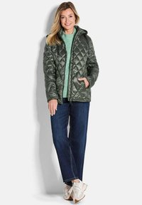 Grüne gepolsterte Steppjacke mit Kapuze, Reißverschluss vorne, seitlichen Taschen, getragen über einem hellgrünen Pullover und dunkelblauen Jeans, kombiniert mit hellen Sneakers.