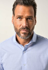 Uomo con capelli corti scuri e barba sale e pepe, che indossa una camicia a quadri azzurri chiari e guarda dritto davanti a sé.