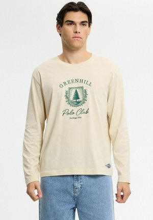 Joven con camisa de manga larga beige con texto verde "Greenhill Polo Club" y emblema de árbol, combinado con vaqueros azul claro.