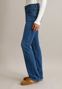 Blaue, ausgestellte Jeans aus Denim mit mittlerer Taille, fünf Taschen und leichten Nähten, kombiniert mit hellbraunen Schnürschuhen.