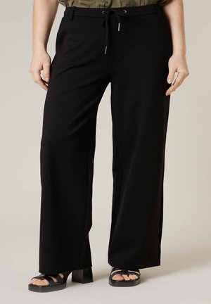 Personne portant un pantalon noir ample avec taille à cordon, associé à des sandales à talons noires sur un fond uni.