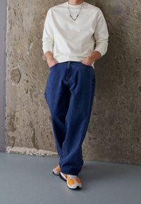 Camisa blanca de manga larga con un pequeño acento amarillo, combinada con jeans azules holgados y zapatillas multicolores con detalles en naranja, blanco y negro.