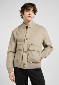 Jonge man in een beige wollen bomberjack met grote zakken en knopen, staat tegen een lichtgrijze achtergrond en kijkt iets opzij.