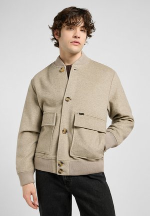 Young man in a beige wool bomber jacket with large pockets and buttons, stands against a light grey background, looking slightly to the side.