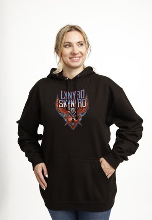 Uśmiechnięta kobieta w czarnej bluzie z kapturem z kolorowym logo Lynyrd Skynyrd, z rękami w kieszeniach, stojąca na białym tle.