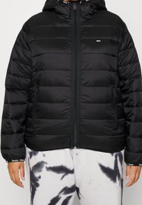 Black puffer jacket with a hood, featuring horizontal stitching, front zip closure, and side pockets. Worn over tie-dye sweatpants.