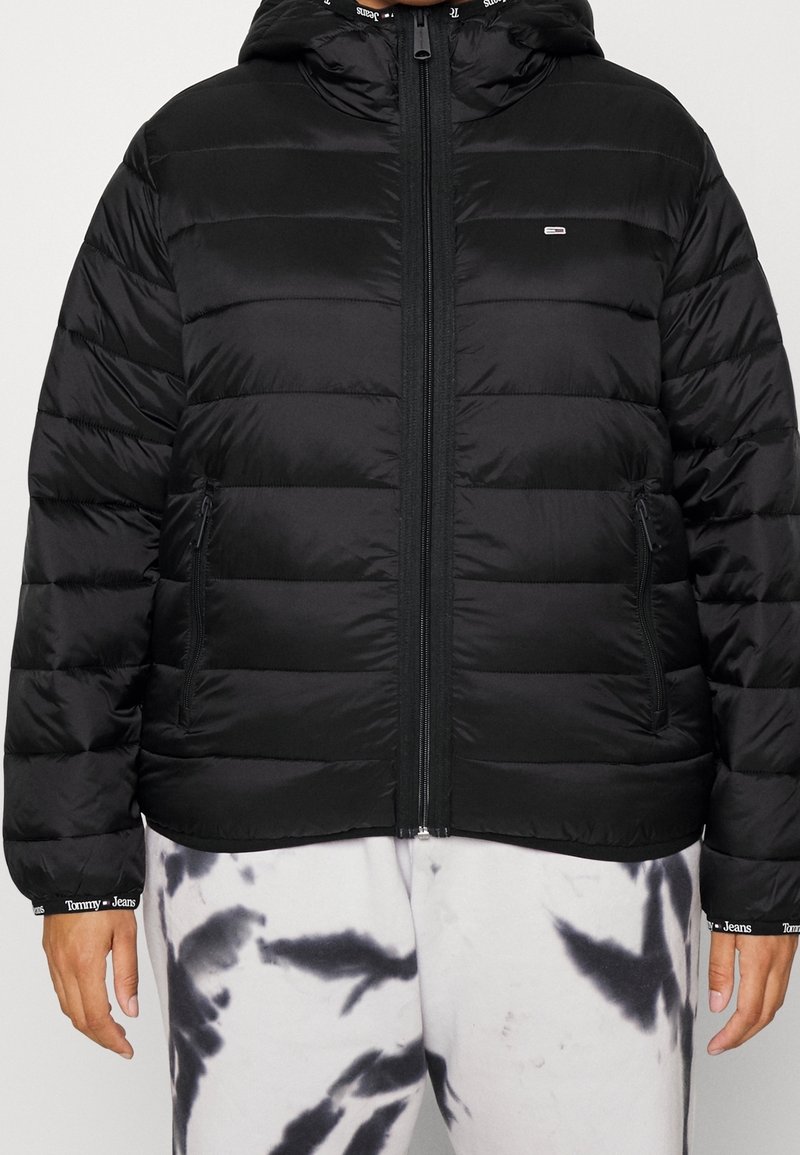 Black puffer jacket with a hood, featuring horizontal stitching, front zip closure, and side pockets. Worn over tie-dye sweatpants.